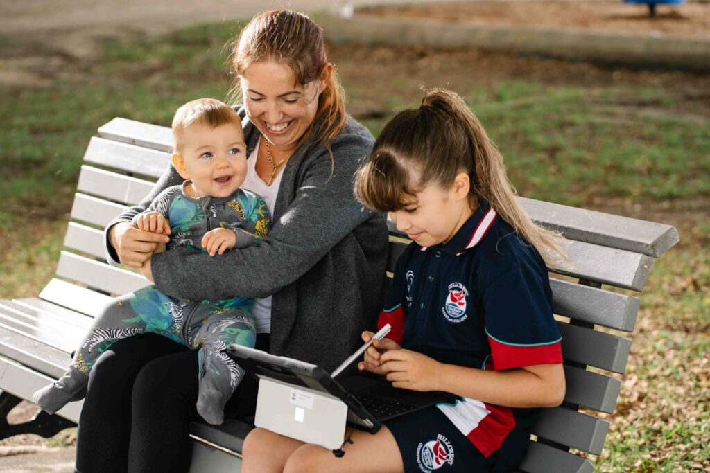 a mother holding her baby chats to a young girl sitting on a park bench studying on her computer